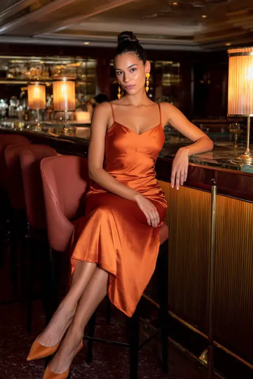A woman in a burnt orange satin midi dress with pointed heels and gold earrings, seated at a chic dinner lounge under warm lighting 