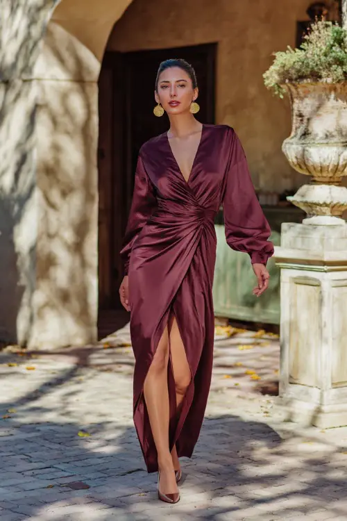 A woman in a deep wine satin wrap maxi dress with a slit, styled with gold earrings and heels, strolling past an elegant fall courtyard
