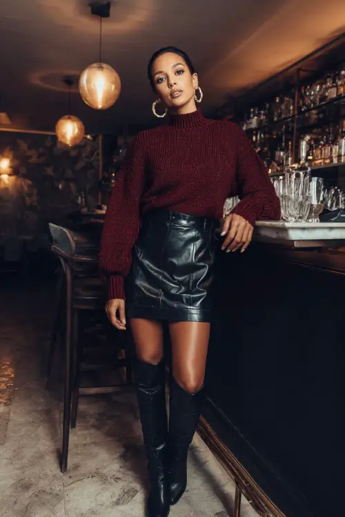 A woman in a maroon sweater tucked into a black leather skirt with tall boots and hoop earrings, standing by a stylish dinner bar 