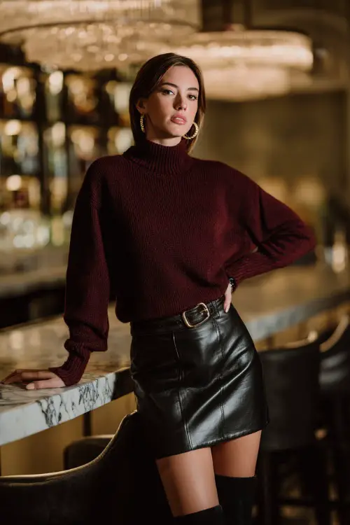 A woman in a maroon sweater tucked into a black leather skirt with tall boots and hoop earrings, standing by a stylish dinner bar 