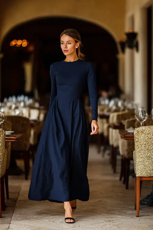 A woman in a navy blue long-sleeve maxi dress with subtle pleats, styled with block heels and pearl earrings, walking into a fine dining restaurant 