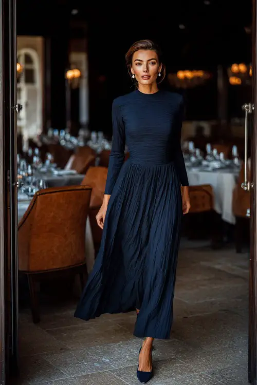 A woman in a navy blue long-sleeve maxi dress with subtle pleats, styled with block heels and pearl earrings