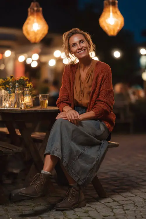 A woman over 40 in a denim midi skirt with a rust cardigan and ankle boots, enjoying a casual dinner outdoors under glowing fall lanterns 
