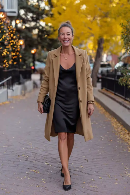 A woman over 50 in a chic camel coat layered over a black satin slip dress, styled with block heels and a clutch, walking along a cozy autumn street with golden leaves 