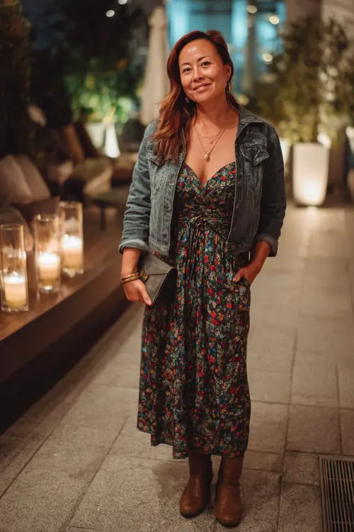 A woman over 50 in a floral wrap midi dress with a denim jacket and ankle boots, holding a clutch while standing near a candlelit outdoor terrace
