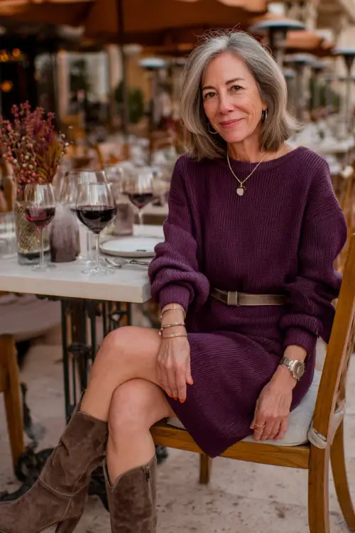 A woman over 50 in a plum-colored knit sweater dress cinched with a belt, paired with suede boots and delicate jewelry, dining at an outdoor terrace with fall décor 