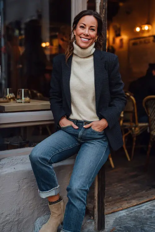 A woman over 50 in dark blue jeans with a cream knit turtleneck, accessorized with a tailored blazer and ankle boots, smiling outside a cozy fall dinner café