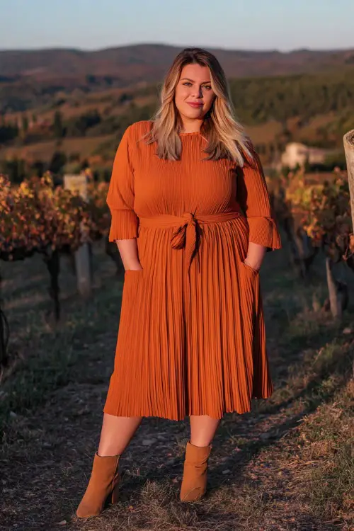 A plus-size woman wears a rust-orange pleated midi dress with long sleeves and a tie belt, paired with tan ankle boots