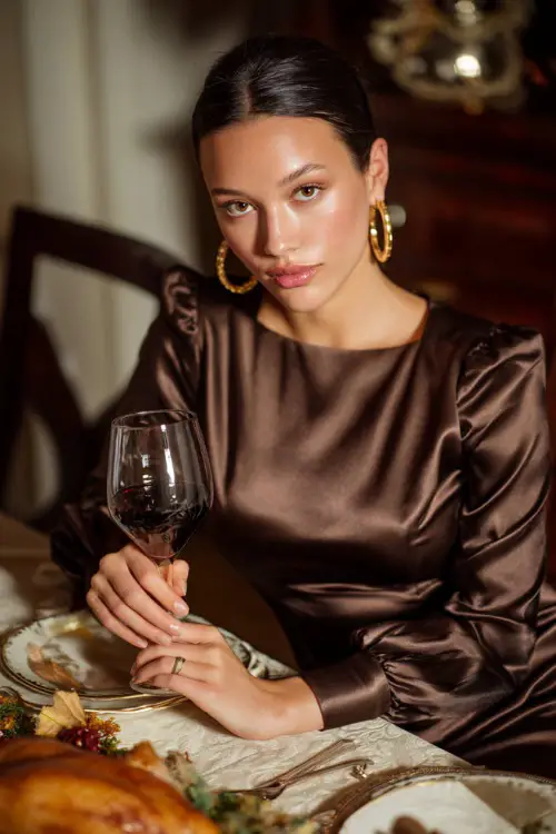 A woman in a chocolate brown satin midi dress with long sleeves, gold hoop earrings, and heels, holding a glass of wine at a Thanksgiving dinner