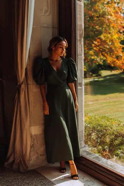 A woman in a dark green maxi dress with puff sleeves and heeled sandals, standing by a window with fall foliage outside