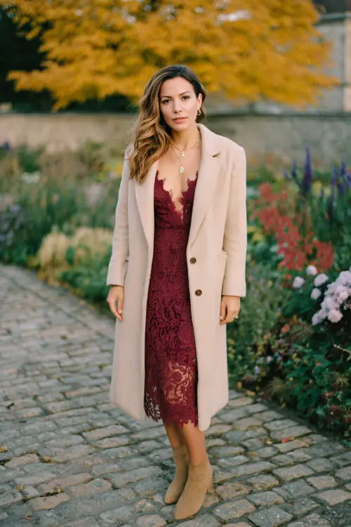A woman over 30 wears a burgundy lace midi dress paired with a long beige wool coat, standing on a cobblestone path surrounded by autumn flowers and golden leaves 