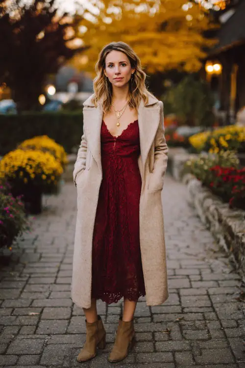A woman over 30 wears a burgundy lace midi dress paired with a long beige wool coat, standing on a cobblestone path surrounded by autumn flowers and golden leaves