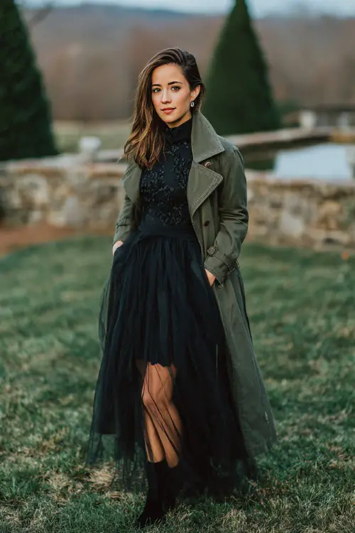 A woman wears a black long-sleeve tulle dress with embroidered details, styled with heeled boots and a dark green trench coat