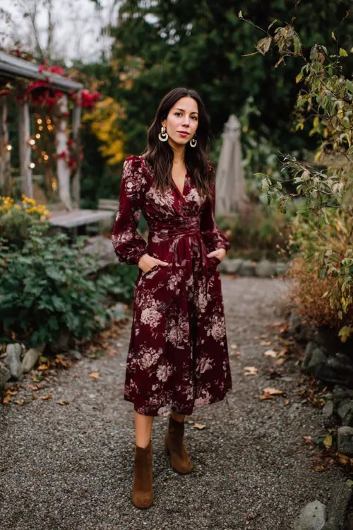 A woman wears a burgundy floral midi dress with long sleeves, paired with suede ankle boots and hoop earrings, walking through a cozy outdoor November wedding garden