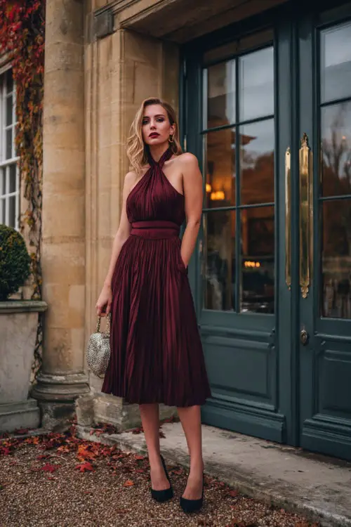 A woman wears a burgundy pleated chiffon dress with a halter neckline and matching belt, paired with black pumps and a metallic handbag 
