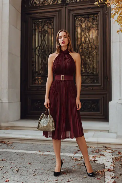 A woman wears a burgundy pleated chiffon dress with a halter neckline and matching belt, paired with black pumps and a metallic handbag