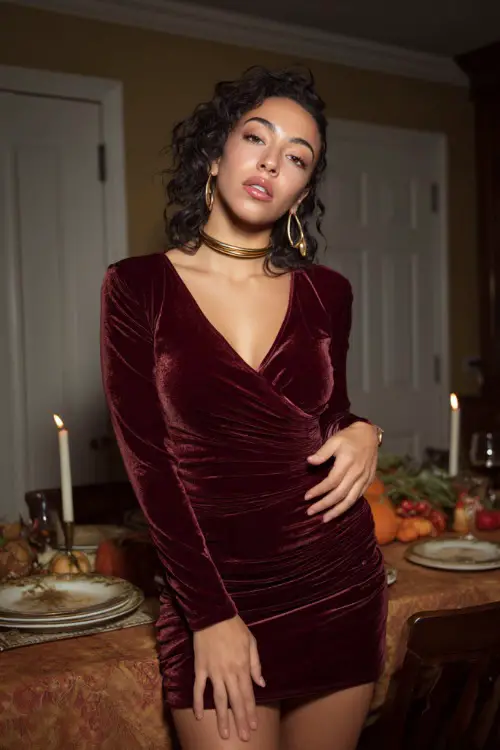 A woman wears a burgundy velvet mini dress with long sleeves and gold accessories, posing near a softly lit Thanksgiving dinner setup