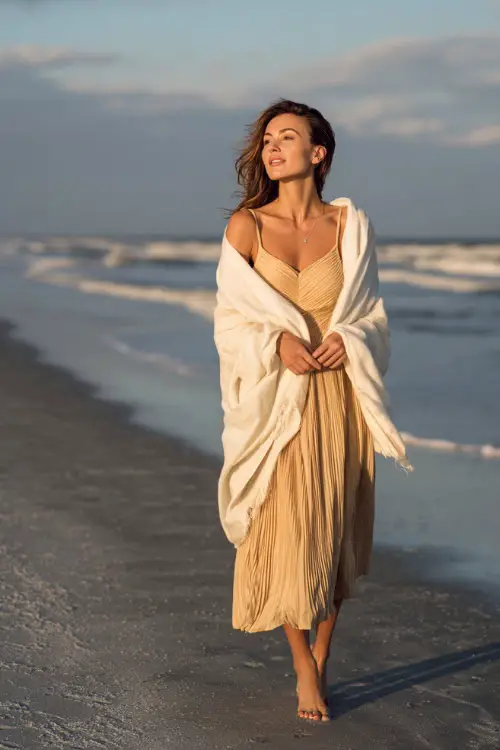 A woman wears a champagne pleated midi dress with thin straps, layered with a soft white shawl