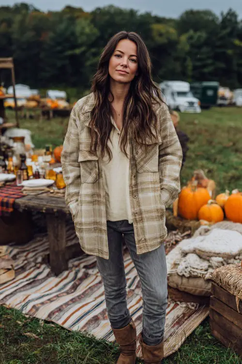 A woman wears a checked shacket over a cream top and jeans, standing near a rustic outdoor Thanksgiving picnic setup with pumpkins and blankets