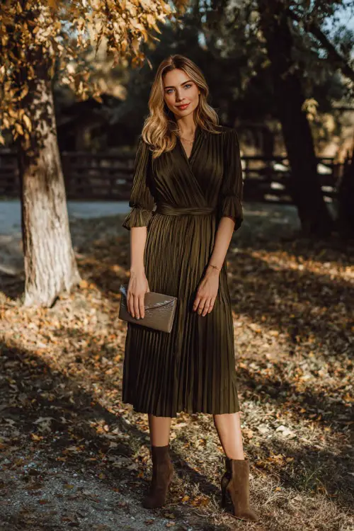 A woman wears a dark olive green pleated midi dress with a belted waist, paired with suede ankle boots and a clutch 
