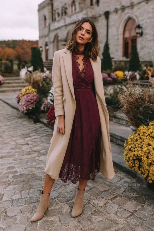A woman wears a deep wine-colored midi dress with lace sleeves, layered with a light tan overcoat and suede booties 