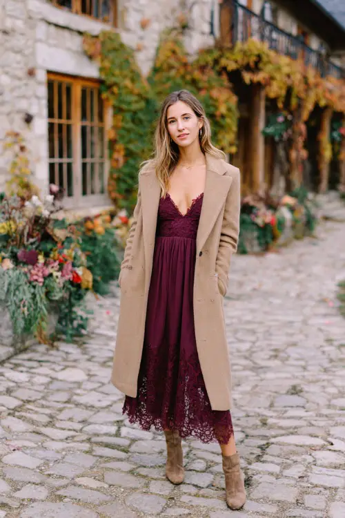 A woman wears a deep wine-colored midi dress with lace sleeves, layered with a light tan overcoat and suede booties