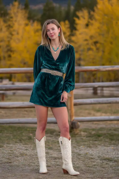 A woman wears a forest green velvet dress with long sleeves and a belted waist, styled with cream cowgirl boots and turquoise jewelry 