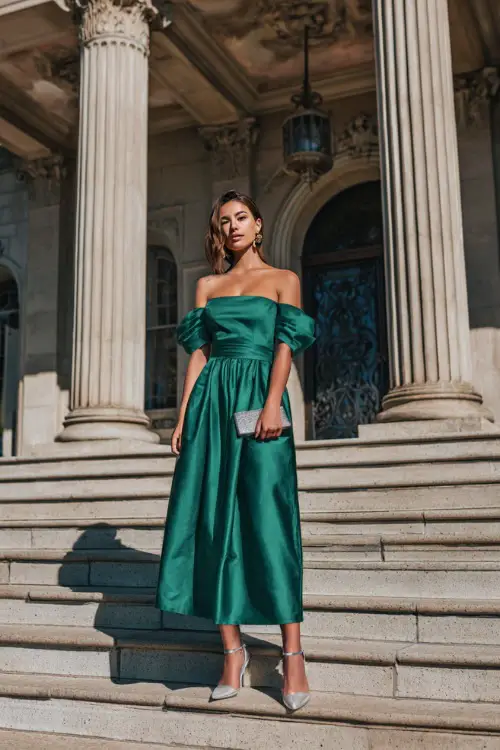A woman wears a jade green off-the-shoulder gown with a fitted bodice and flowy skirt, accessorized with silver sandals and a matching clutch 