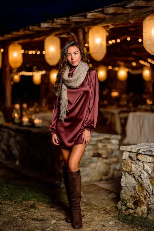 A woman wears a maroon long-sleeve satin dress with a high neckline, paired with knee-high boots and a wool scarf