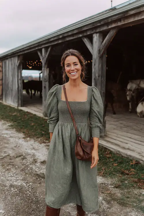 A woman wears a moss green midi dress with puff sleeves and a square neckline, styled with brown boots and a leather crossbody bag, posing at a casual barn wedding in November