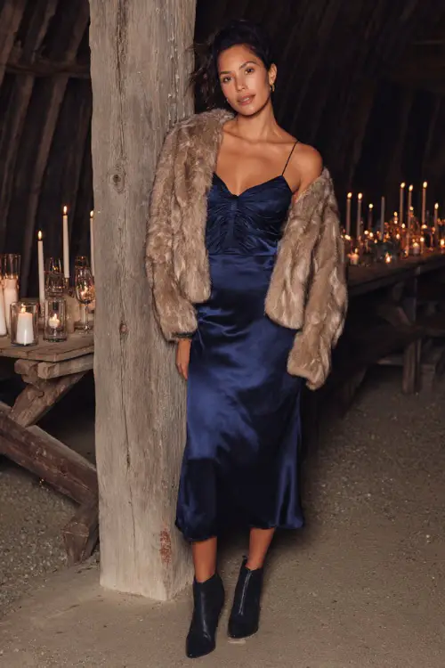 A woman wears a navy blue satin midi dress with puff sleeves, styled with a cropped faux fur jacket and ankle boots, posing at a candlelit wedding reception in a rustic barn