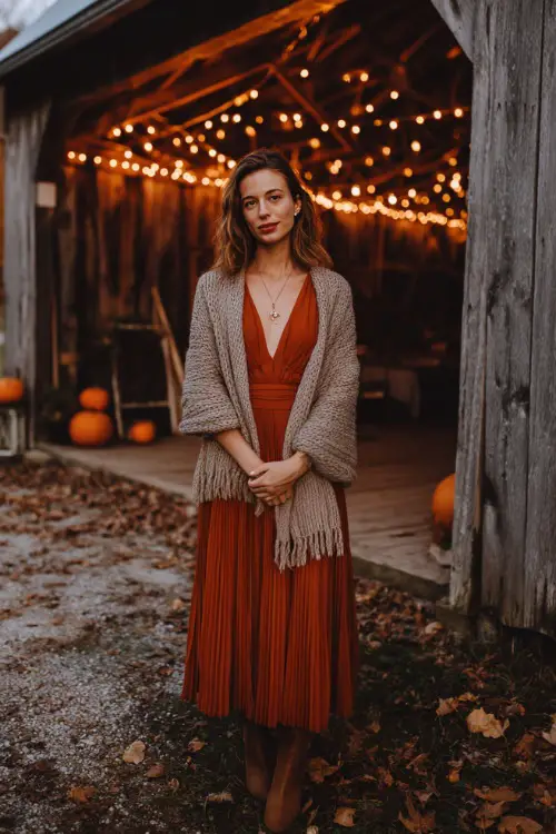 A woman wears a pleated rust-orange midi dress with a chunky knit taupe wrap around her shoulders 