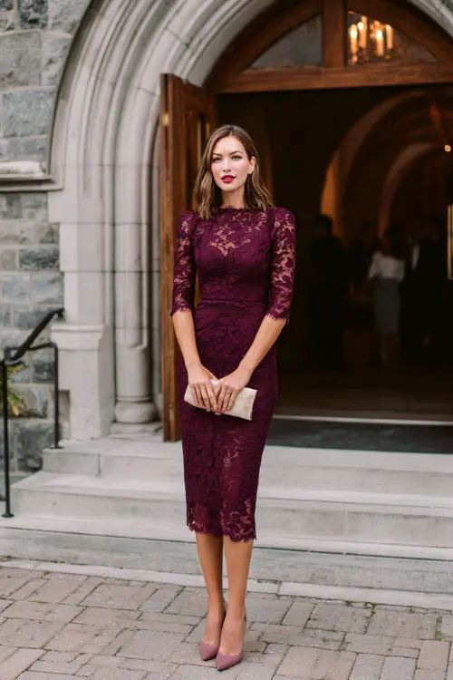 A woman wears a plum lace midi dress with three-quarter sleeves and a scalloped hemline, paired with pointed-toe heels and a clutch