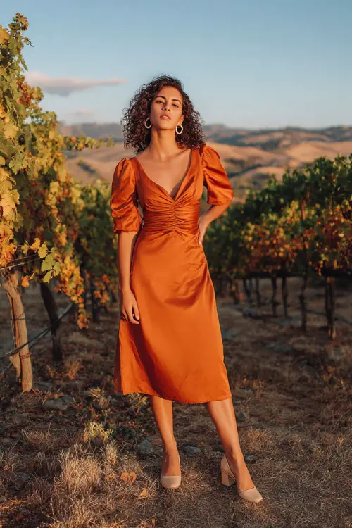 A woman wears a rust-orange satin midi dress with a ruched bodice and flutter sleeves, paired with beige block heels and gold hoops 
