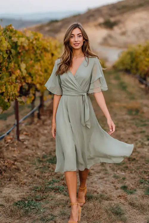 A woman wears a sage green chiffon midi dress with flutter sleeves and a tie waist, paired with tan block heels 