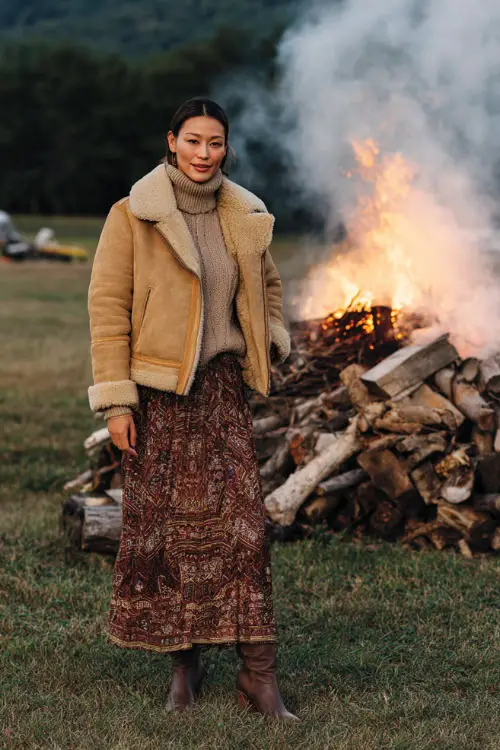 A woman wears a tan shearling jacket over a sweater and maxi skirt with heeled boots