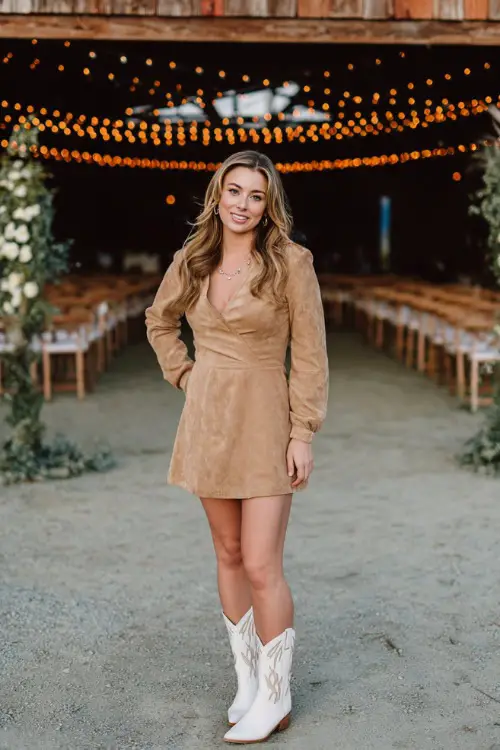 A woman wears a tan suede dress with long sleeves and a fitted waist, styled with white cowboy boots and silver jewelry