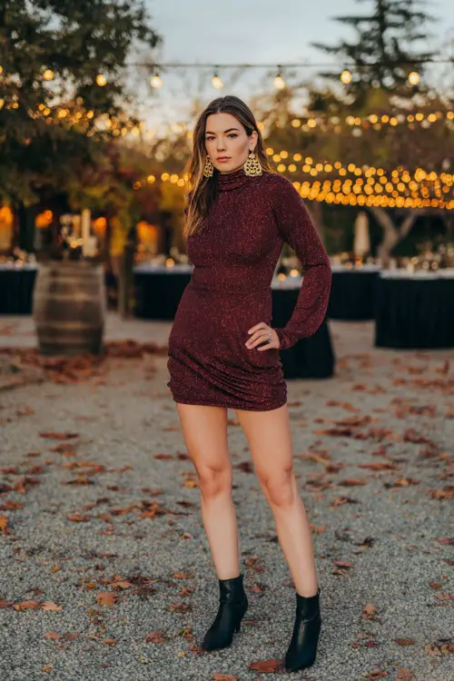 A woman wears a wine-red long-sleeve mini dress with a high neckline and subtle shimmer, paired with black heeled boots and gold earrings 
