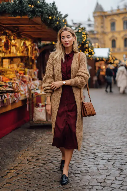 A woman in a camel sweater layered over a burgundy midi shirt dress, gold watch, leather crossbody bag, and black heeled loafers, walking through a refined European Christmas street market 