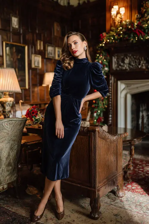 A woman in a navy velvet high-neck dress with puff sleeves, diamond earrings, and heeled Mary Janes, posing inside a stately home with festive garlands and rich wooden furniture