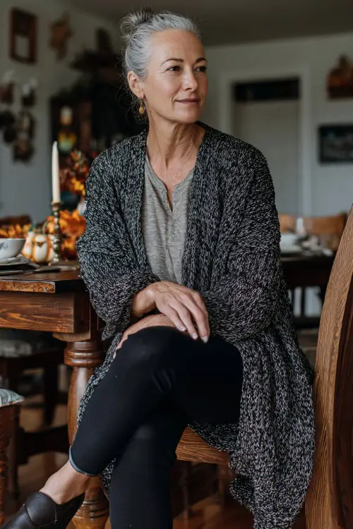 A woman over 50 in a long cardigan over a soft top and black leggings, wearing loafers while chatting with family near a Thanksgiving dining table