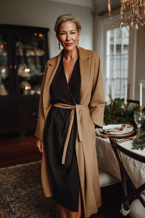 A woman over 50 wears a belted camel dress coat over a black inner knit set, pearl studs, leather heels, full body framing in a softly styled Christmas dinner table setting at home 