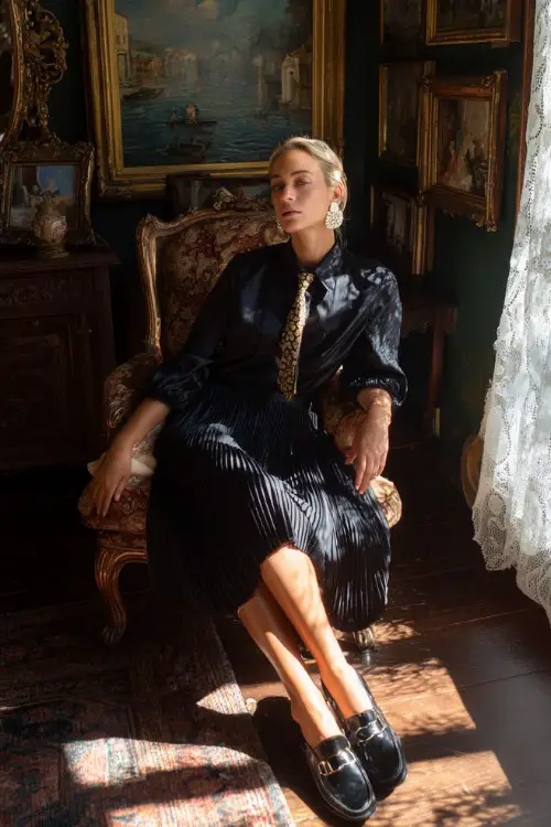 A woman over 50 wears a navy pleated dress with a silk necktie, heeled loafers, and pearl drop earrings