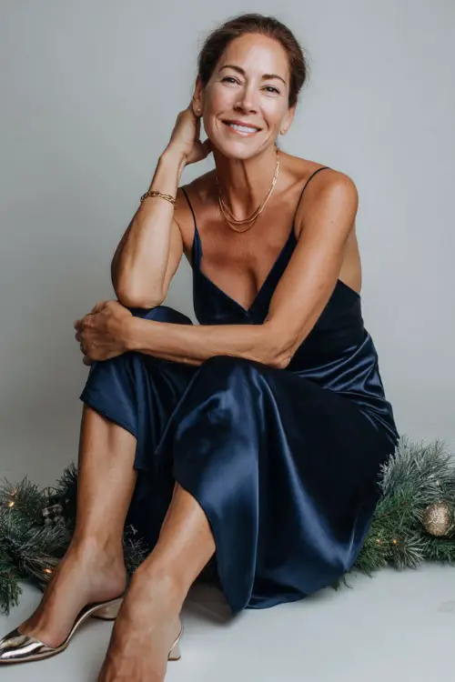A woman over 50 wears a navy satin midi gown, delicate gold bracelet, metallic pointed heels, full body shot in a minimalist studio with a soft Christmas garland on the floor side 