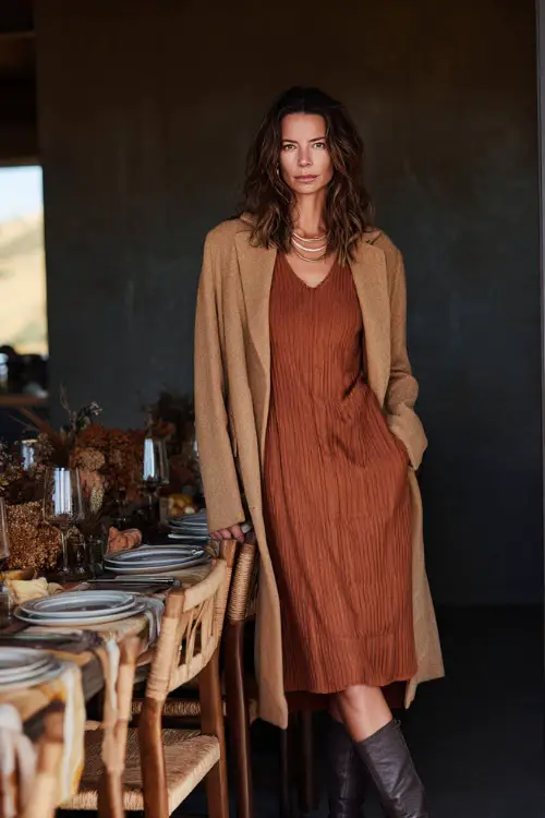 A woman over 50 wears a rust-colored knit dress with a long camel coat and heeled boots, standing beside a beautifully set Thanksgiving dining table