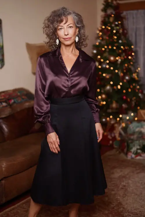 A woman over 50 wears a satin plum blouse tucked into a black A-line midi skirt, pearl drop earrings, low heels