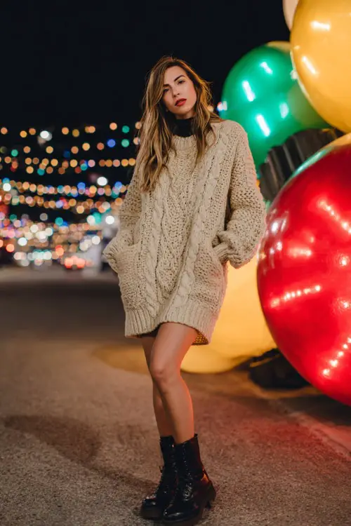 A woman wearing a chunky knit sweater dress, long coat layer, leather boots, full-body visible, standing beside oversized Christmas light ornaments