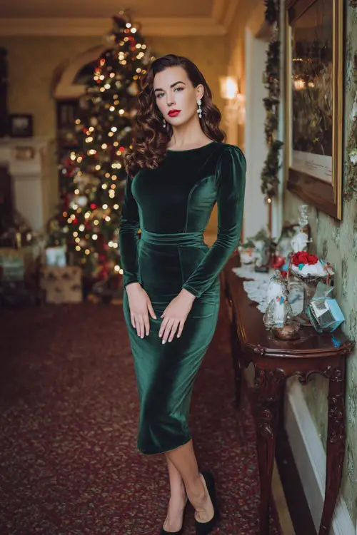 A woman wearing a deep green velvet midi dress with a fitted waist, pearl earrings, classic black heels, and soft curls, standing in an elegant Christmas-decorated mansion 