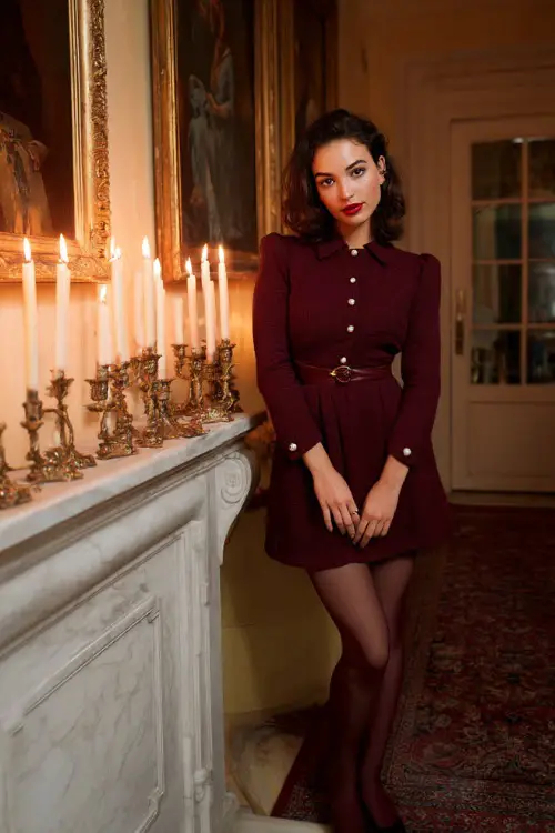 A woman wearing a tailored burgundy tweed dress with a cinched waist, pearl studs, sheer tights, and Mary Jane heels, standing in a classic European living room glowing with holiday candles 