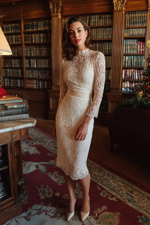 A woman wearing an ivory lace midi dress with scalloped edges, diamond studs, and polished pumps, standing in a refined library with classic Christmas décor